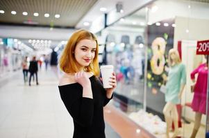 moda garota ruiva usar vestido preto com make brilhante segurando a xícara de café no centro comercial. filtros de instagram de estilo de foto em tons.