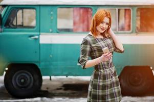 senhora jovem ruiva com telefone celular móvel e fones de ouvido, vestindo no vestido xadrez fundo velho ônibus de minivan turquesa vintage. foto