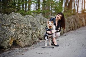 menina cigana morena com cachorro yorkshire terrier posou contra pedras no parque. modelo usa jaqueta de couro e camiseta com enfeite, calça e sapatos com salto alto. foto