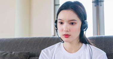 mulher feliz asiática estudante usar fones de ouvido estudar on-line assistindo podcast de webinar no laptop ouvindo aprendizagem curso de educação telefonando e escrevendo notas olhe para o laptop sentado no sofá em casa