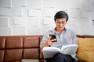 homem asiático sênior lendo livro e usando telefone inteligente no sofá na sala de estar em casa retrato de homem idoso asiático é relaxante e felicidade com ler uma revista foto