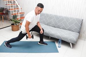 homem asiático de camisa branca no tapete azul levantando peso com mini halteres em casa foto
