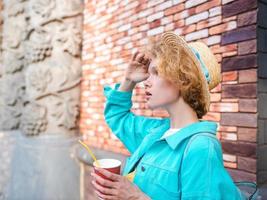 jovem ruiva encaracolada alegre na jaqueta jeans azul e chapéu de palha segurando a xícara de café e surpreso com pontos de referência. diversão, verão, viagens, moda, conceito de juventude foto