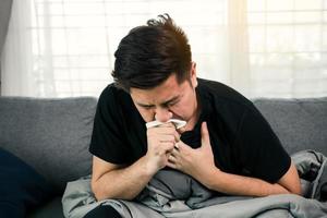 os asiáticos estão doentes ou doentes com bronquite enquanto tossem, cobrindo a boca com papel de seda quando ele se senta no sofá em casa.