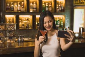 mulher asiática tirando uma foto de si mesma enquanto bebe uísque no bar.