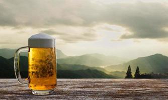 chope ou cerveja artesanal em vidro transparente com espuma de cerveja em cima, coloque no conjunto de mesas e cadeiras de madeira. sala de estar no terraço de madeira com fundo de montanha com luz de Deus ou renderização em 3d foto