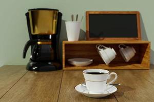 café preto em uma caneca de cerâmica branca com aro e pires dourados. borrão máquina de café expresso preto e dourado e armazenamento de xícara de café em fundo branco e papel de parede branco. mesa de madeira e verde suave foto