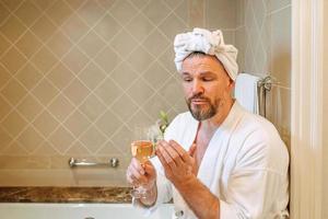 homem maduro bonito em roupão branco e toalha na cabeça sentado no banheiro com espuma e bebendo vinho rosé. spa, relax, estilo de vida, aproveite o conceito de vida. foto