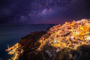 vila de oia na luz do sol com estrelas e via láctea, santorini, grécia. incrível paisagem de férias de verão, arquitetura branca e luzes noturnas. destino de viagem famoso, fundo de viagens urbanas foto