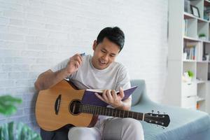 homem asiático tocando violão enquanto está sentado no sofá na sala de luz. homem asiático escrevendo música enquanto toca guitarra em casa. compor o conceito de música de música. foto