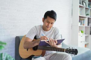 homem asiático tocando violão enquanto está sentado no sofá na sala de luz. homem asiático escrevendo música enquanto toca guitarra em casa. compor o conceito de música de música. foto