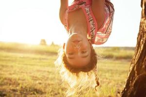 a garota está pendurada de cabeça para baixo em uma árvore no verão na luz do sol laranja e um vestido leve. horário de verão, calor, infância. retrato engraçado com cabelo desgrenhado foto
