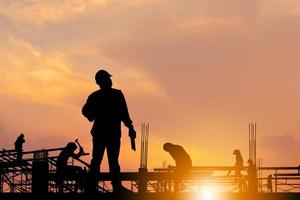 silhueta de engenheiro e trabalhador verificando projeto no fundo do canteiro de obras, canteiro de obras ao pôr do sol na noite. foto