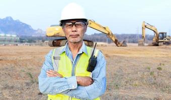 engenheiro masculino sênior asiático vestindo colete de segurança e capacete inspeciona obras civis o engenheiro-chefe de construção usa um walkie-talkie para gerenciar a escavadeira. foto