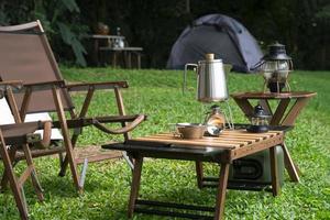 chaleira de aço inoxidável, fogão a gás portátil, tigela e lanternas vintage com mesa ao ar livre em gramado verde na área de camping foto