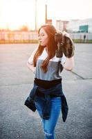 jovem adolescente urbana com skate, usar óculos, boné e jeans rasgados no pátio de esportes no pôr do sol. foto