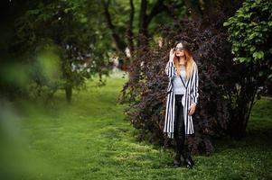 mulher elegante olha com paletó listrado preto e branco, calça de couro e óculos de sol posando contra arbustos na rua. conceito de menina da moda. foto