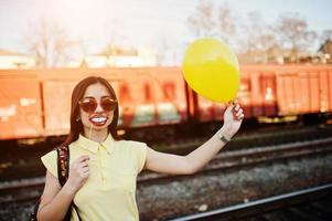jovem adolescente em pé na plataforma da estação de trem com lábios engraçados na vara e balão na mão, use camiseta amarela, jeans e óculos de sol, com mochila. foto