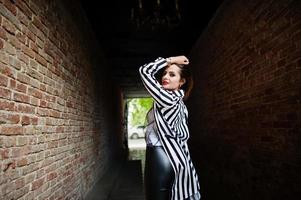 parte de trás do look de mulher elegante com paletó listrado preto e branco, calça de couro, posando no velho túnel de tijolo vermelho da rua. conceito de menina da moda. foto