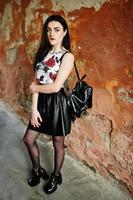 jovem gótica na saia de couro preta com mochila posou contra a parede do grunge. foto