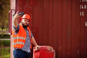 trabalhador de barba homem terno trabalhador da construção civil no capacete laranja de segurança perto do barril vermelho faz selfie no telefone. foto