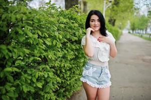 retrato de menina morena sexy em shorts jeans feminino e blusa branca contra arbustos verdes de primavera. foto