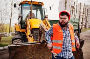 homem trabalhador barba brutal terno trabalhador da construção civil no capacete laranja de segurança, contra traktor com chave ajustável na mão. foto