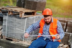 homem trabalhador barba terno trabalhador da construção civil no capacete laranja de segurança contra o pavimento com uma fita métrica na mão. foto