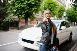 mulher loira elegante usa jeans, óculos e jaqueta de couro com telefone celular, contra carro de luxo. retrato de modelo urbano de moda. foto