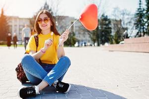 jovem adolescente sentado nas ruas da cidade com balão de pirulito e coração nas mãos, use camiseta amarela, jeans e óculos de sol.