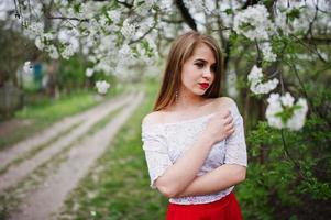 retrato de menina bonita com lábios vermelhos no jardim de flor de primavera, use vestido vermelho e blusa branca. foto