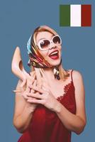 Engraçado elegante sorrindo alegre chique loira mulher com maquiagem e sapato de salto alto nos braços e a bandeira italiana no fundo foto