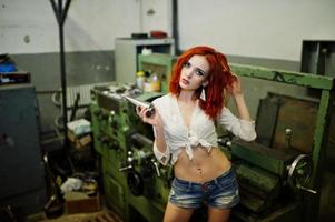 garota ruiva usa shorts jeans curtos e blusa branca posada na máquina industrial na fábrica. foto