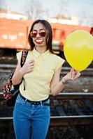 jovem adolescente em pé na plataforma da estação de trem com lábios engraçados na vara e balão na mão, use camiseta amarela, jeans e óculos de sol, com mochila. foto