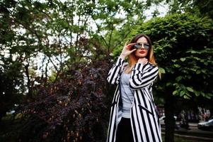 mulher elegante olha com paletó listrado preto e branco, calça de couro e óculos de sol posando contra arbustos na rua. conceito de menina da moda. foto