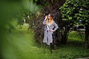 mulher elegante olha com paletó listrado preto e branco, calça de couro e óculos de sol posando contra arbustos na rua. conceito de menina da moda. foto