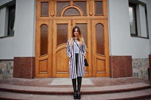 olhar de mulher elegante com paletó listrado preto e branco, calça de couro, posando contra a grande porta de madeira do edifício. conceito de menina da moda. foto