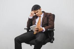 decepcionado empresário asiático de paletó e gravata está sentado e recebe más notícias do telefone inteligente foto