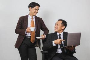 dois empresários asiáticos de terno sorrindo e olhando um ao outro sentado e apontando para o laptop enquanto toma um café foto
