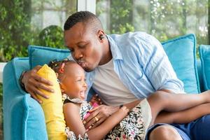 alegre pai e filha afro-americanos brincando na sala de estar, jovem pai beijando bebê alegre, conceitos de família de felicidade foto