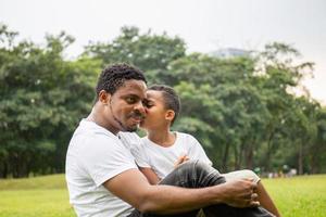 Africano menino bonitinho no pai beijando casual no parque. feliz pai afro-americano enquanto abraça e carrega seu filho sentado ao ar livre, alegre conceito de família negra. foto