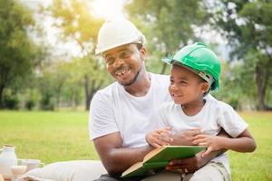 alegre menino afro-americano e pai fazendo um piquenique no parque, feliz filho e pai lendo um livro, conceitos de família de felicidade foto