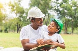 afro-americano pai e filho no capacete lendo um livro, feliz pai e filho fazendo um piquenique no parque, conceitos de família de felicidade foto