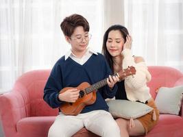 duas mulheres compartilhando amor e tocando ukulele no sofá sofá na sala de estar em casa, se divertindo juntos, casal de lésbicas foto