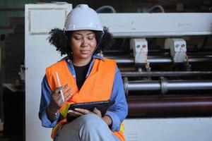 trabalhadora engenheira afro-americana em uniforme de segurança e capacete, inspeção de qualidade por tablet, manutenção, verifique a máquina na indústria de fabricação de produção de papel, gerente técnico de fábrica. foto