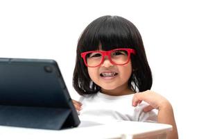 feliz menina pré-escolar asiática usando óculos vermelhos e usando tablet pc em fundo branco e sorriso, menina asiática aprendendo com uma chamada de vídeo com tablet, conceito educacional para crianças em idade escolar foto