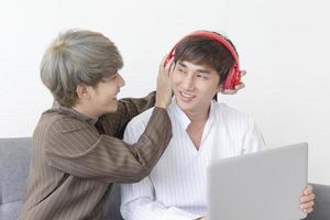 um casal masculino com um homem asiático sentado em um sofá com seu amor um pelo outro e outro ouvindo música tocando seu laptop. foto