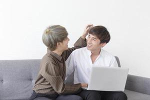 um casal masculino com um homem asiático sentado em um sofá com seu amor um pelo outro e outro jogando laptop. foto