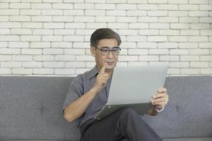 um homem asiático de meia-idade segurando um notebook na mão está sentado em um sofá fazendo uma chamada de vídeo. foto