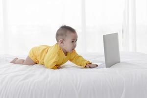 um bebê asiático está brincando em um laptop em uma cama branca e limpa. foto